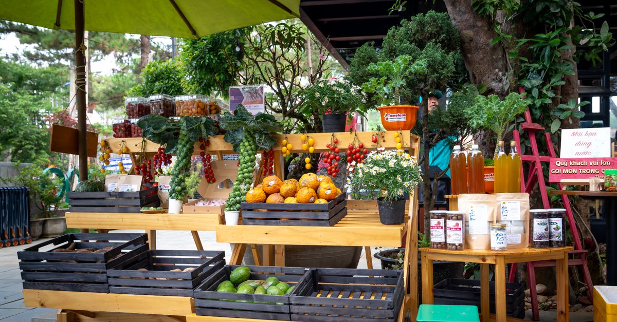 A colorful outdoor market stand showcasing fresh fruits, vegetables, and products under a green umbrella.